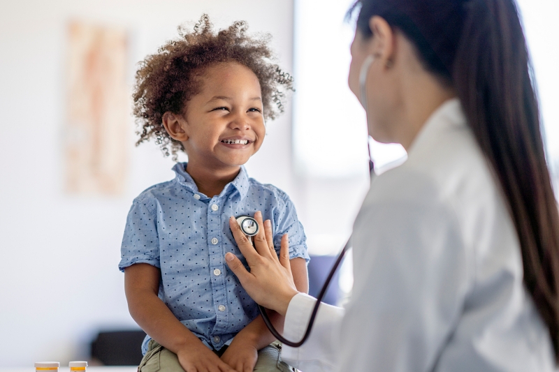 A female doctor of Asian decent listens to her patients heart with her stethoscope. The little boy is sitting up on the exam table smiling back at his doctor as she holds the instrument to his chest.